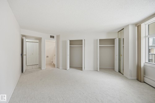 Light-colored carpeting throughout, featuring two open closet spaces, a mirrored closet door, and a large window with a balcony visible - 504 11710 100 Avenue, Edmonton, AB 