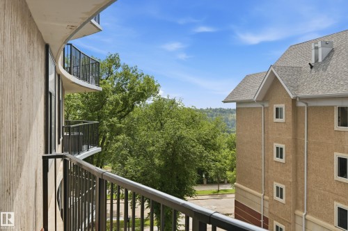 Private balcony with black metal railing and textured wall, overlooking mature trees and a residential building with gabled roof lines - 504 11710 100 Avenue, Edmonton, AB 