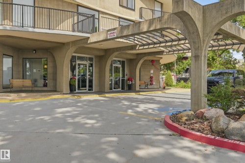 Building entrance featuring a covered drive-up area with a concrete pergola structure, multiple glass entrance doors, and an exterior balcony with black railings - 504 11710 100 Avenue, Edmonton, AB 