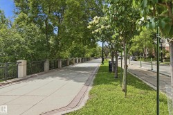 Paved pedestrian walkway featuring a brick border, metal railing with stone posts, and mature deciduous trees - 