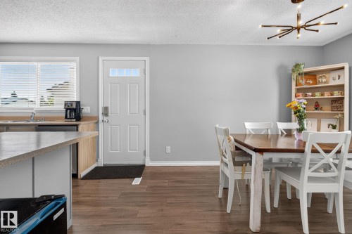Open-concept kitchen and dining area featuring wood-finish flooring, a kitchen island with a light-toned countertop, a window with horizontal blinds, a white paneled entry door with clear glass inserts, and a contemporary ceiling light fixture - 3218 41 Street, Leduc, AB 