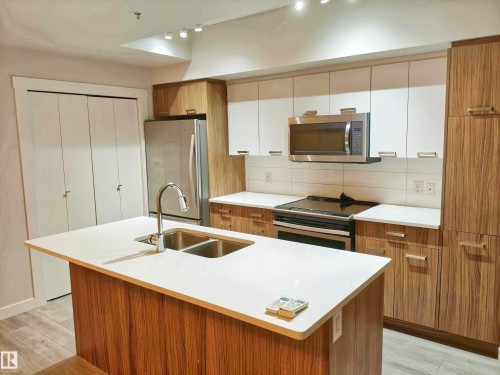 Kitchen featuring an expansive island with a double basin sink, stainless steel appliances, two-tone cabinetry with wood-finish and white panels, white countertops, and wood-finish flooring - 2505 10410 102 Avenue, Edmonton, AB 