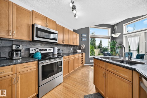 Kitchen featuring light wood cabinetry, stainless steel appliances, dark countertops, a dark tile backsplash, and wood-finish flooring - 8507 179 Avenue, Edmonton, AB 