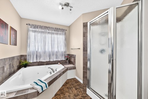 Spacious bathroom featuring an integrated corner tub with tile surround, a separate glass-enclosed shower, a window with privacy sheers, track lighting, and tile-finish flooring - 8507 179 Avenue, Edmonton, AB 