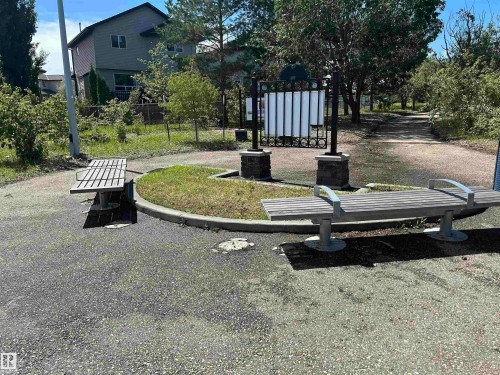 Paved outdoor area featuring two slatted benches with metal armrests, a central decorative sign structure with stone bases, and a natural grass pathway beyond - 8507 179 Avenue, Edmonton, AB 