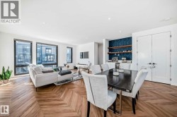 Open-concept living and dining area featuring chevron-patterned wood-finish flooring, a built-in wet bar with wood shelving and a patterned accent wall, multiple large windows with dark frames, and double white panel doors - 