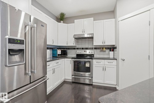 U-shaped kitchen featuring white cabinetry, stainless steel appliances including a refrigerator and range, a tile backsplash with a decorative accent, dark countertops, and dark wood-finish flooring - 4320 Annett Common, Edmonton, AB - Indoor Photo Showing Kitchen