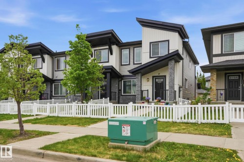 Modern townhome exterior featuring an asymmetrical roofline, light-toned vertical siding, dark grey horizontal siding, stone-clad columns, and a white picket fence - 4320 Annett Common, Edmonton, AB - Outdoor