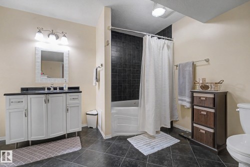 Bathroom featuring dark tile flooring, a white vanity with a dark countertop, a white bathtub with dark tile surround, a wall-mounted mirror with a decorative frame, and a chrome light fixture - 19039 47 Avenue, Edmonton, AB 