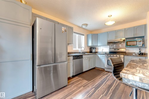 Kitchen featuring light blue cabinetry, stainless steel appliances, wood-finish flooring, and a window with blinds - 312 Beringer Crescent, Edmonton, AB 