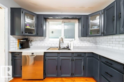 Kitchen featuring dark cabinetry with glass-front upper cabinets, white subway tile backsplash, light-toned countertops, and a stainless steel dishwasher - 20 Chippewa Rd, Leduc, AB 