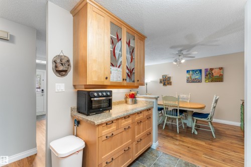 Kitchenette with light wood-finish cabinetry, frosted glass cabinet doors, and a speckled countertop, opening to a dining area with wood-finish flooring and a ceiling fan - 5802 172 Street, Edmonton, AB 