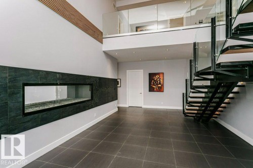 Two-story space featuring a linear fireplace with dark tile surround, a floating spiral staircase with glass railing, an upper-level glass railing, wood-finish wall accents, and large format floor tile - 9035 Saskatchewan Drive, Edmonton, AB 
