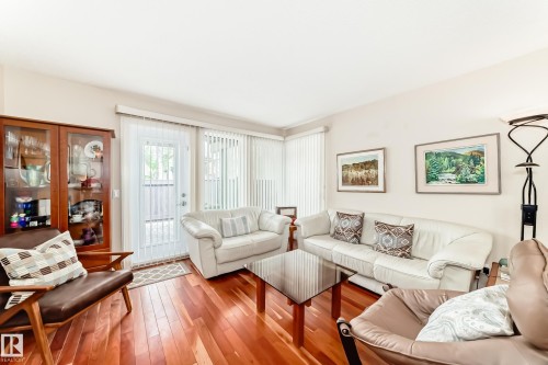Living room featuring rich wood-finish flooring, light-toned walls, and abundant natural light from vertical blinds, gas fireplace, and patio door access to private patio. - 7930 111 Street, Edmonton, AB - Indoor Photo Showing Living Room