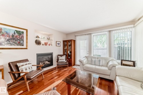 Living area featuring wood-finish flooring, a built-in fireplace, recessed shelving with glass accents, and large windows with vertical blinds - 7930 111 Street, Edmonton, AB - Indoor Photo Showing Living Room With Fireplace