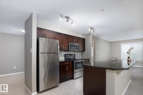 Kitchen featuring a dark wood cabinetry, stainless steel appliances, a dark countertop island with an integrated double sink, and light wood-finish flooring - 102 25 Element Dr, St. Albert, AB 