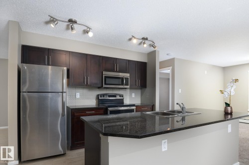 Kitchen featuring dark wood cabinetry, stainless steel appliances, a tile backsplash, and a breakfast bar with a double basin sink - 102 25 Element Dr, St. Albert, AB 