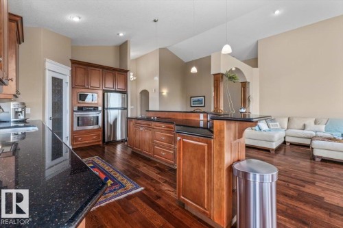 Gourmet kitchen featuring rich wood cabinetry, stainless steel appliances, dark stone countertops, wood-finish flooring, and a vaulted ceiling - 215 Westerra Point(E), Stony Plain, AB 
