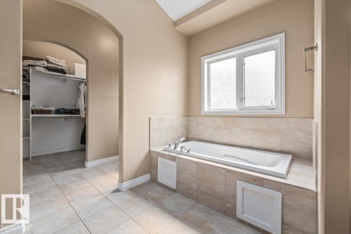 Soaking tub with tile surround and white window, complemented by large format floor tiles and an arched doorway leading to a walk-in closet with built-in shelving - 215 Westerra Point(E), Stony Plain, AB 
