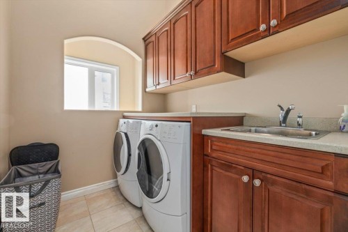 Laundry area featuring wood-finish cabinetry, a stainless steel utility sink, composite countertop, and tiled flooring - 215 Westerra Point(E), Stony Plain, AB 