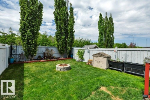 Fenced backyard featuring a stone-look fire pit, green lawn, and tall privacy trees - 6042 214 Street, Edmonton, AB - Outdoor