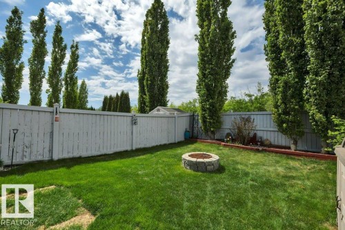 Expansive backyard featuring a stone-ringed fire pit, manicured lawn, tall privacy trees, and a wooden perimeter fence - 6042 214 Street, Edmonton, AB - Outdoor