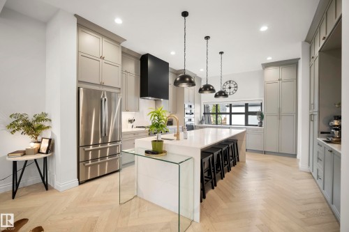Contemporary kitchen featuring light gray cabinetry, stainless steel appliances, a large center island with seating, pendant lighting, and light wood-finish flooring in a herringbone pattern - 6115 111 Avenue, Edmonton, AB - Indoor Photo Showing Kitchen With Upgraded Kitchen