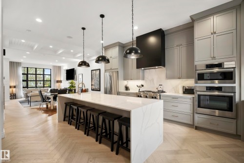 Modern kitchen featuring a waterfall island with integrated seating, light gray cabinetry, stainless steel appliances, a black range hood, and wood-finish herringbone flooring - 6115 111 Avenue, Edmonton, AB - Indoor Photo Showing Kitchen With Upgraded Kitchen