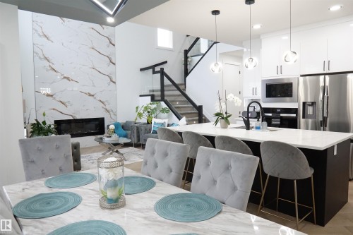 Open-concept living space featuring a floor-to-ceiling fireplace with large format marble-look tile, a modern kitchen island with seating, a glass-panel staircase, and sleek pendant lighting - 17703 74 Street, Edmonton, AB 