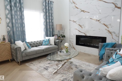 Living area featuring a modern electric fireplace insert set into a large-format porcelain slab accent wall, light wood-finish flooring in a herringbone pattern, and a window with full-length drapery - 17703 74 Street, Edmonton, AB 