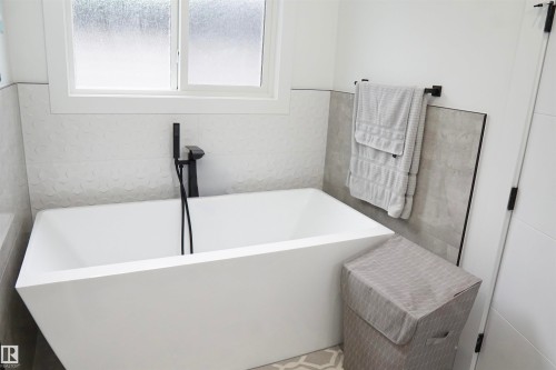 Freestanding soaking tub featuring matte black fixtures, textured white tile surround, and a frosted window - 17703 74 Street, Edmonton, AB 