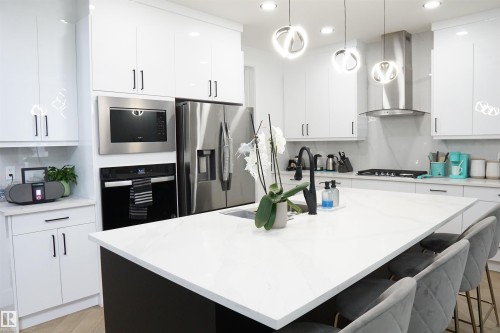 Contemporary kitchen featuring white high-gloss cabinetry, stainless steel appliances, a center island with seating, pendant lighting, and a tile backsplash - 17703 74 Street, Edmonton, AB 