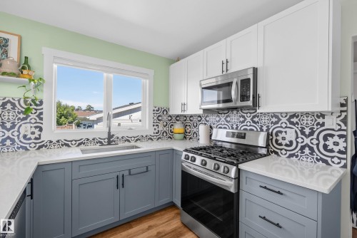 Kitchen featuring two-tone cabinetry with white upper and blue-grey lower cabinets, stainless steel appliances, patterned tile backsplash, and light-toned countertops - 14128 73 Street, Edmonton, AB 