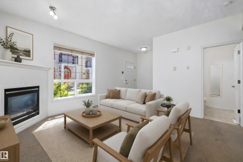 Bright living area featuring a corner fireplace with a white mantle, a large window, and carpet flooring - 101 13825 155 Avenue, Edmonton, AB 