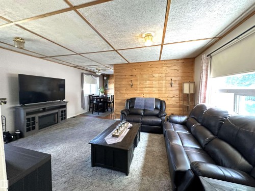 Living area featuring textured ceiling panels with wood-finish trim, a wood-paneled accent wall, and a large window with roller shades - 825 2 Street, Thorhild, AB 