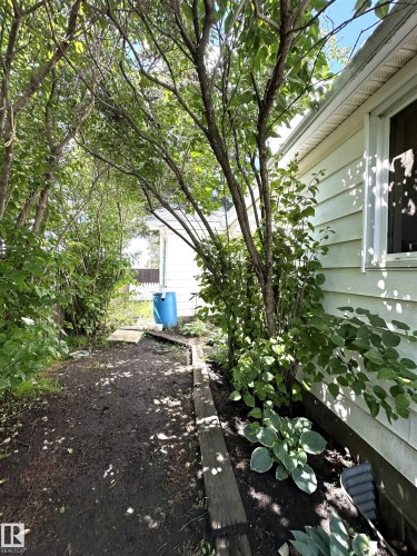 Side yard featuring mature trees, established hosta plants, wood plank edging, and a vinyl-sided exterior - 825 2 Street, Thorhild, AB 