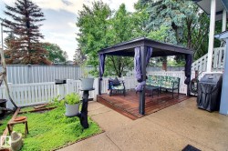 Outdoor living space featuring a dark wood-finish deck with a black gazebo, a white picket fence, a concrete patio, and mature perimeter trees - 