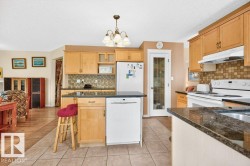 Open-concept kitchen featuring light wood cabinetry, dark granite-style countertops, a tiled backsplash, and integrated white appliances - 