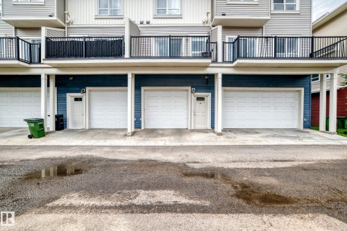 Multiple individual garages with white panel doors and separate entry doors below a multi-level structure featuring balconies with black metal railings - 37 13003 132 Avenue Nw, Edmonton, AB 