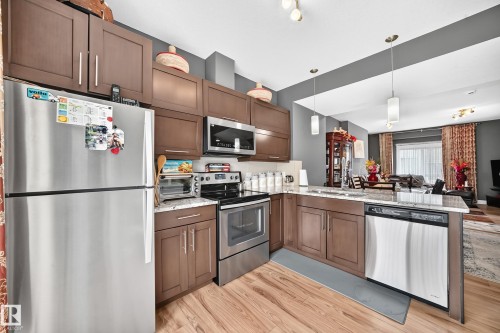 Open-concept kitchen featuring wood-finish flooring, stainless steel appliances, stone-finish countertops, dark wood cabinetry, and pendant lighting - 37 13003 132 Avenue Nw, Edmonton, AB 