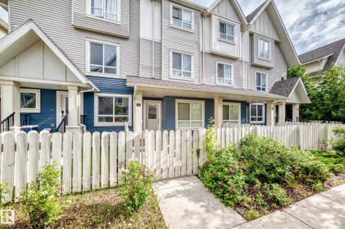 Townhome exterior featuring a white picket fence, a mature landscaped garden, blue and grey siding, multiple windows, and a covered front porch - 37 13003 132 Avenue Nw, Edmonton, AB 