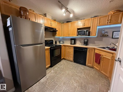 Kitchen featuring ample wood-finish cabinetry, stainless steel refrigerator, black range, and black dishwasher - 205 3425 19 Street, Edmonton, AB 