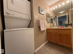 Laundry area with a stacked washer and dryer, adjacent to a vanity featuring wood-finish cabinetry, a white basin sink, and a large mirror with strip lighting - 