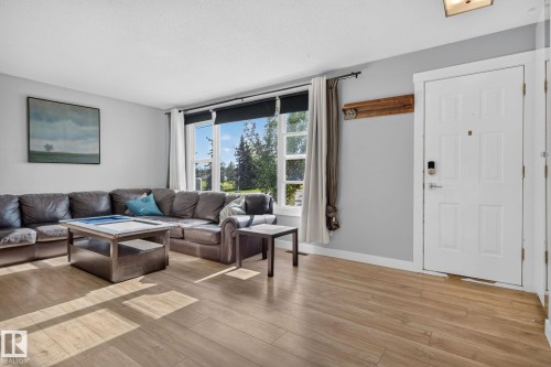 Open-concept living area featuring wood-finish flooring, a large picture window with outdoor scenery, light gray wall paint, and a white entry door with a digital lock - 7803 73 Street, Edmonton, AB 