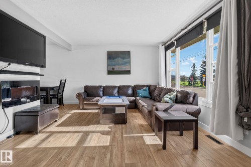 Living area featuring wood-finish flooring, a large window with white framing, and a wall-mounted television above a built-in electric fireplace unit - 7803 73 Street, Edmonton, AB 
