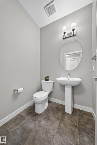 Powder room featuring a pedestal sink, round mirror with a two-light vanity fixture, a toilet, and large format tile flooring - 2331 Casey Crescent Sw, Edmonton, AB 