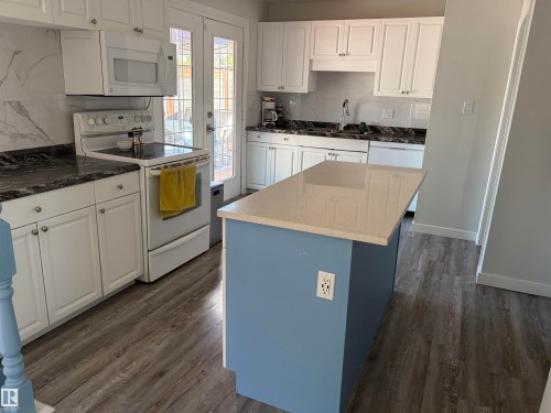 Kitchen featuring white cabinetry, dark stone countertops, a light-colored island with a solid surface countertop, wood-finish flooring, and a white tile backsplash - 15922 92 Avenue, Edmonton, AB 
