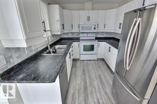U-shaped kitchen with white cabinetry, dark countertops, white subway tile backsplash, stainless steel refrigerator, and wood-finish flooring - 9457 156 Street, Edmonton, AB 