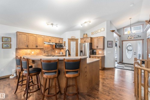 This kitchen features wood cabinetry, a large island with seating, stainless steel appliances, and a tiled backsplash - 61 Linkside Blvd, Spruce Grove, AB 