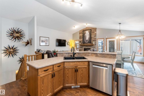 Well-appointed kitchen featuring wood cabinetry, a double basin sink with a black faucet, and a stainless steel dishwasher, with views of the living area with a stone fireplace and vaulted ceilings - 61 Linkside Blvd, Spruce Grove, AB 